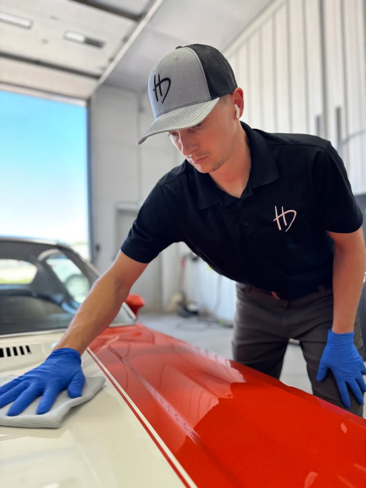 A Hermsmeyer Detailing technician in a branded HD shirt hand-polishing a classic car's hood