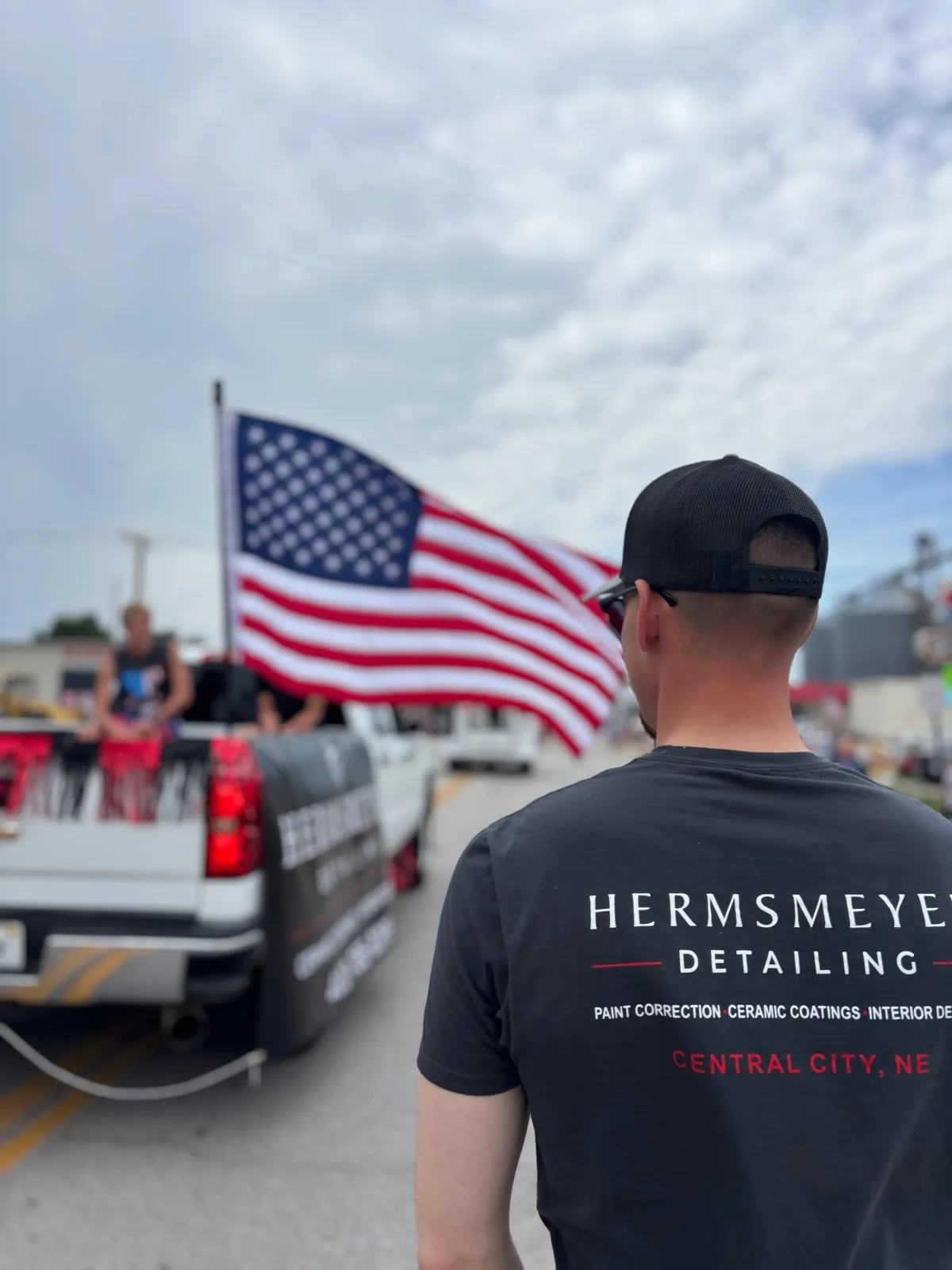 A Hermsmeyer Detailing team member in a branded shirt watching a local parade with the Hermsmeyer Detailing truck and an American flag
